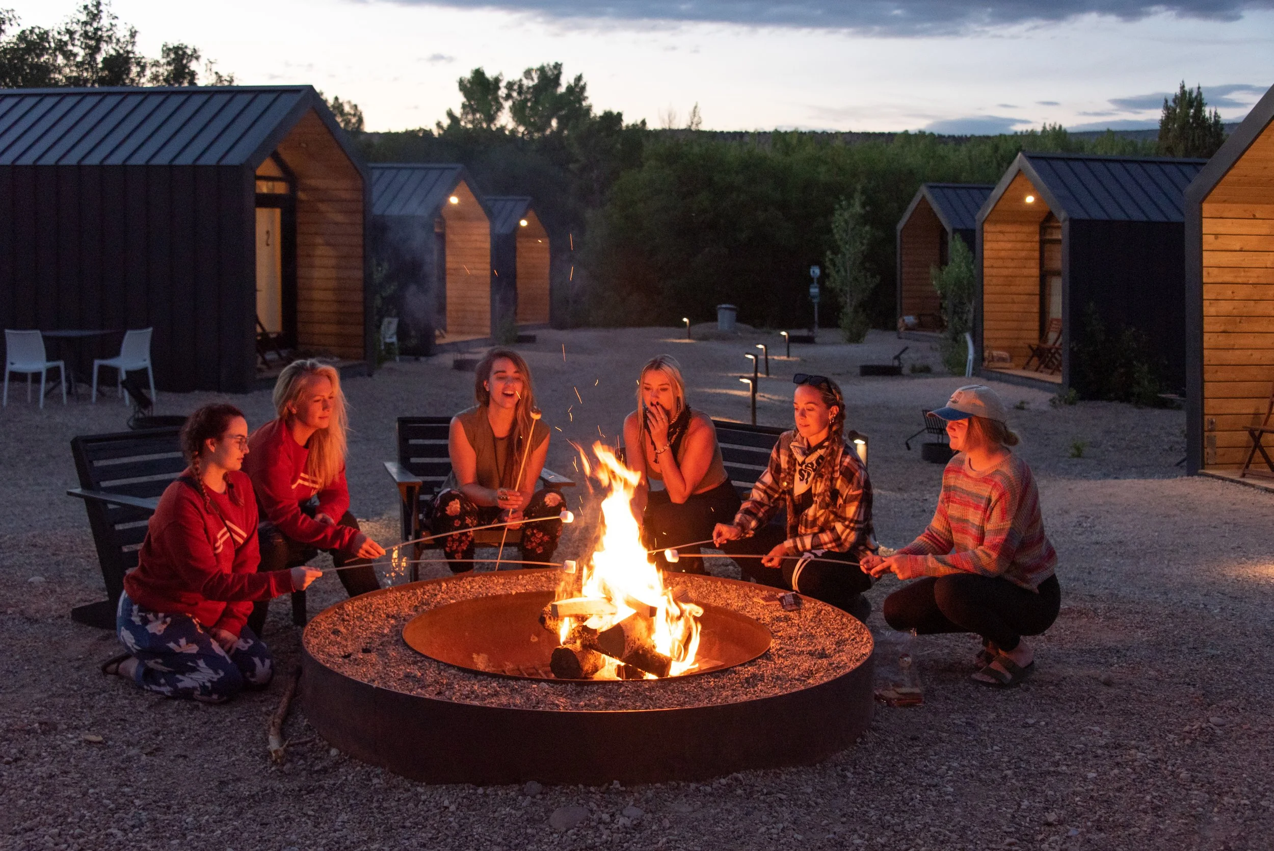 Group of people sitting around a campfire at dusk outside cabins, smiling and enjoying each other's company. Ofland group friendly