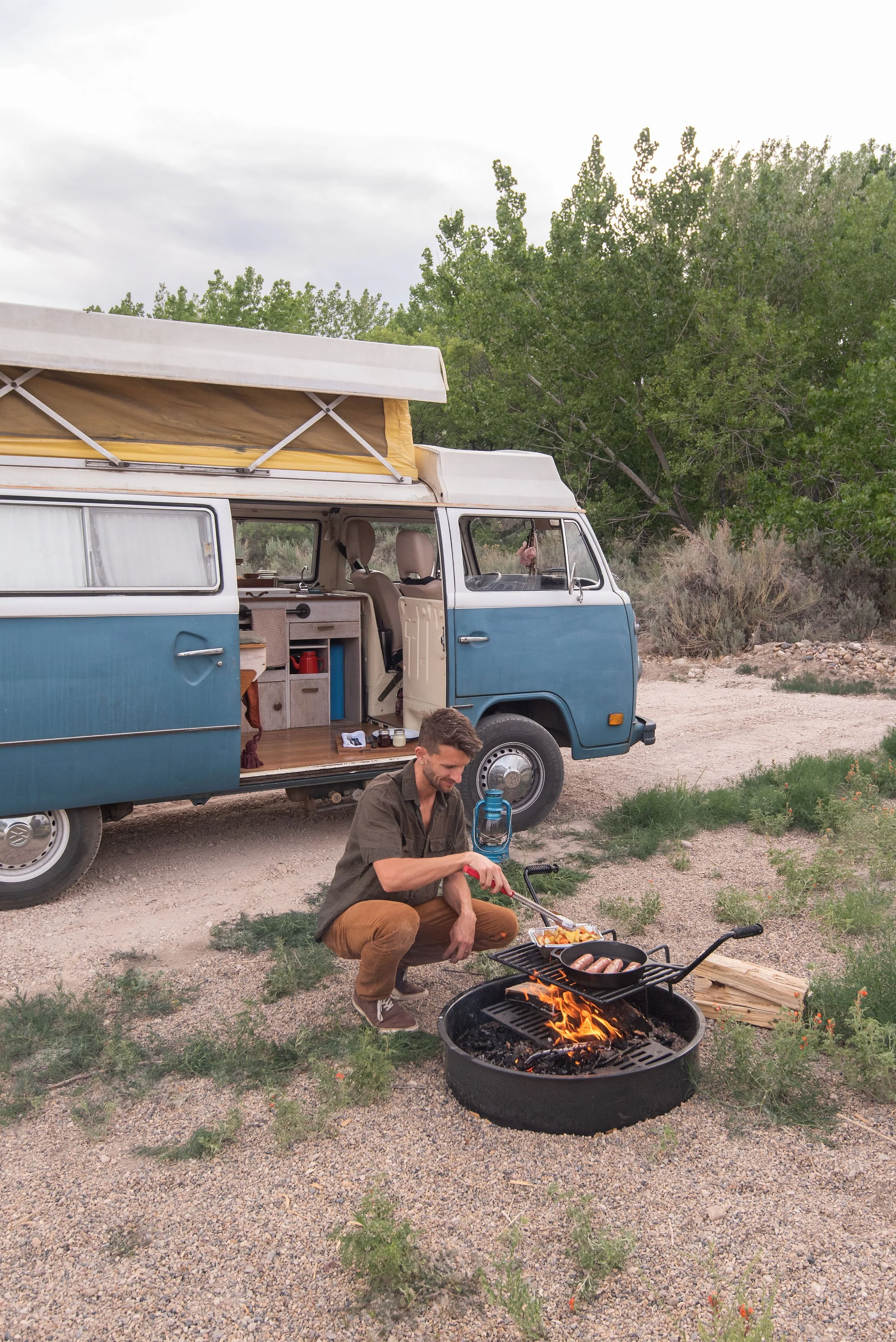 A man cooking hot dogs and vegetables over an open fire grill outside a blue camper van with greenery and trees in the background.