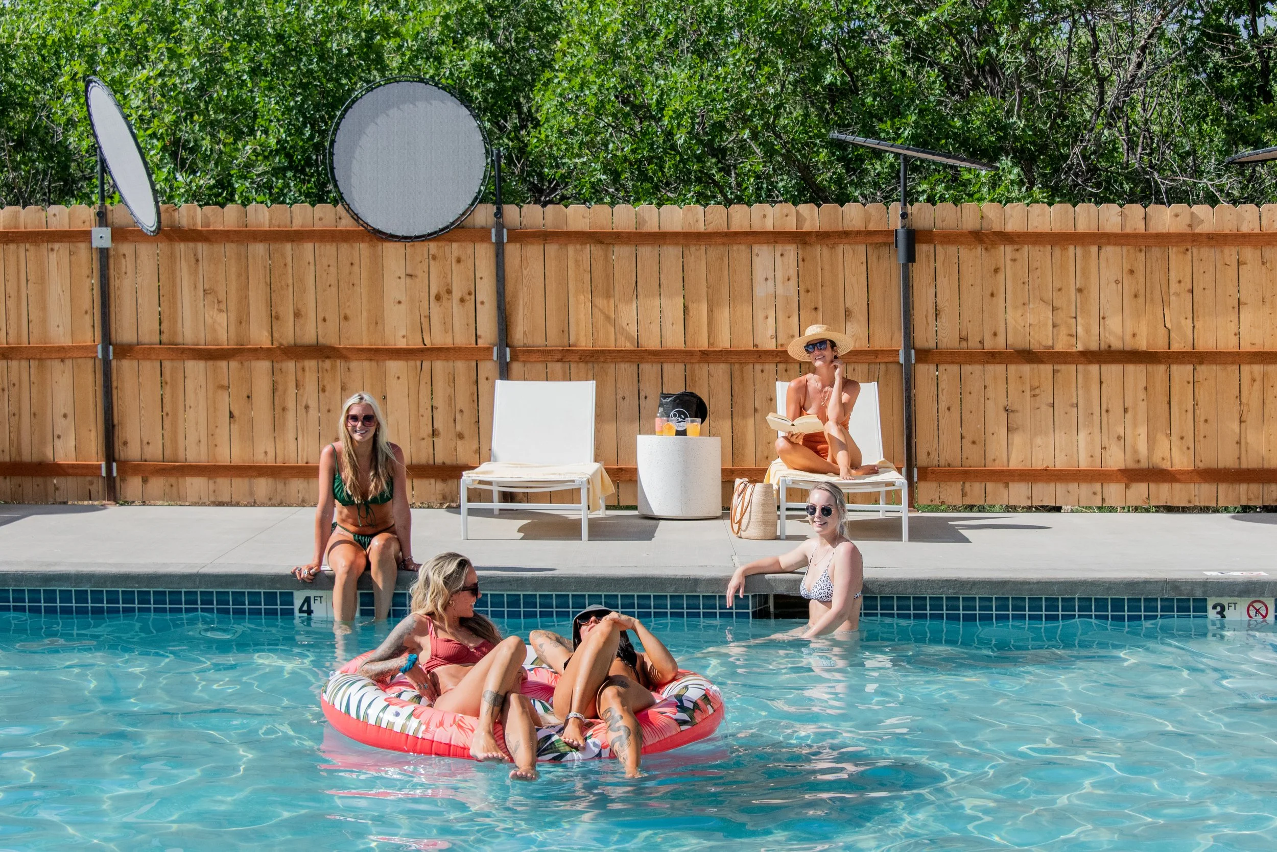Group of women relaxing by a swimming pool, some sitting on the pool edge, others on a float, with a wooden fence and green trees in the background. One woman is reading a book, another is talking on the phone, and several are enjoying the water.