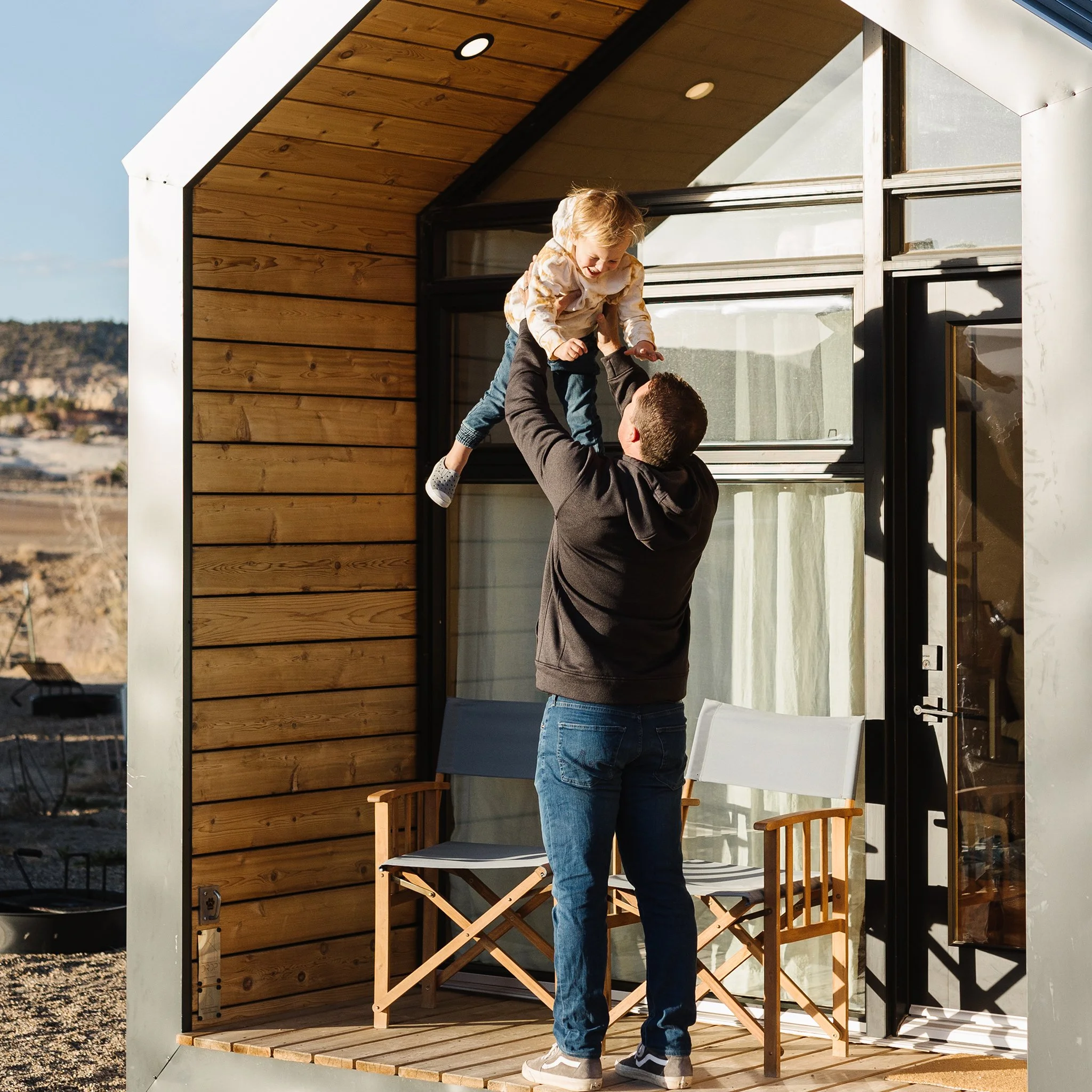 A man lifting a young boy inside a small modern outdoor structure with wooden panel walls and large glass windows, during daytime with hills and a clear sky in the background.