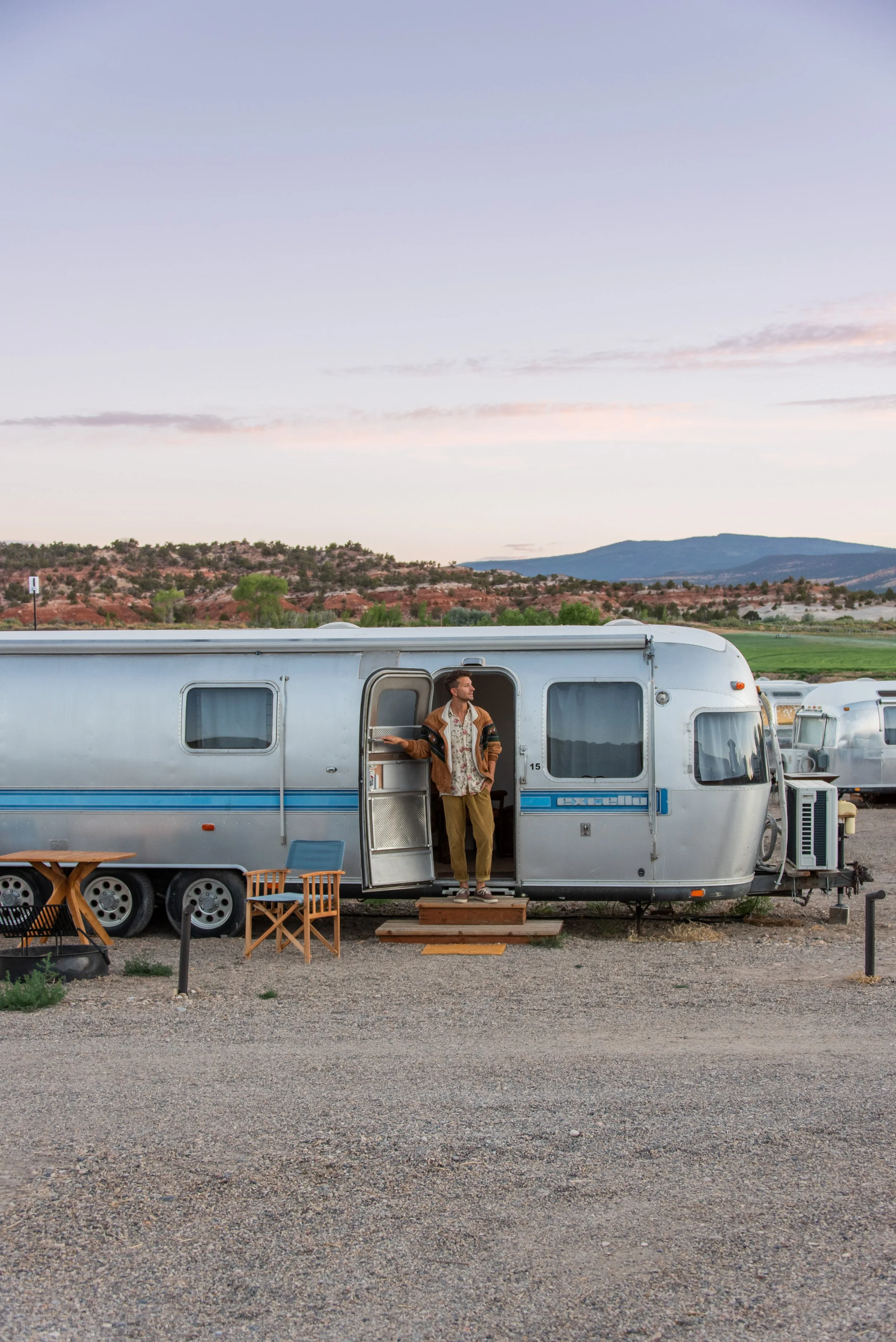 Vintage Airstream - A man stands in the doorway of a vintage Airstream travel trailer with mountains and a pastel sky in the background.