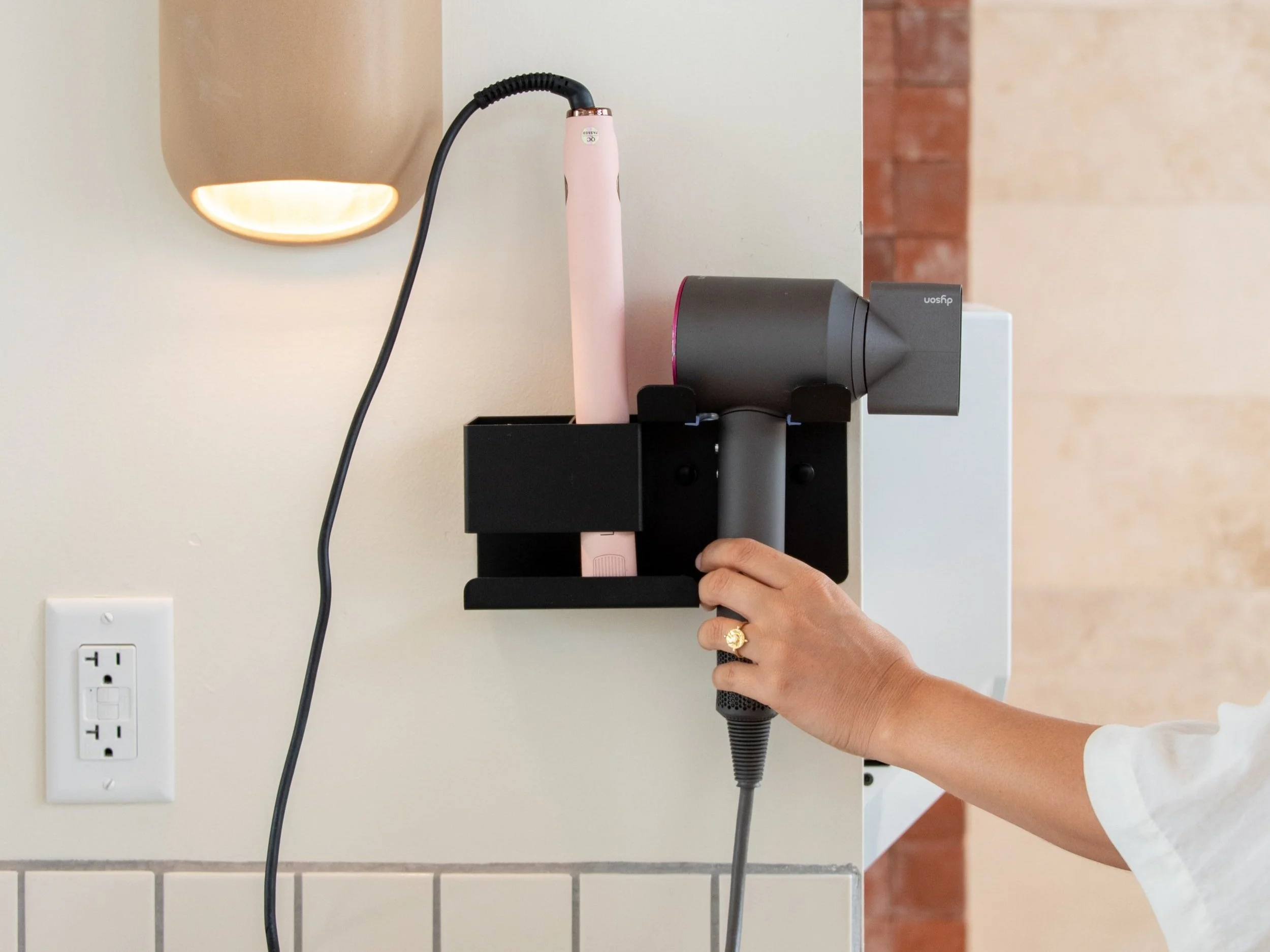 Spa-Quality Bathhouses - A person plugging a black Dyson hairdryer and pink straightener into a wall-mounted black holder on a kitchen or bathroom wall.