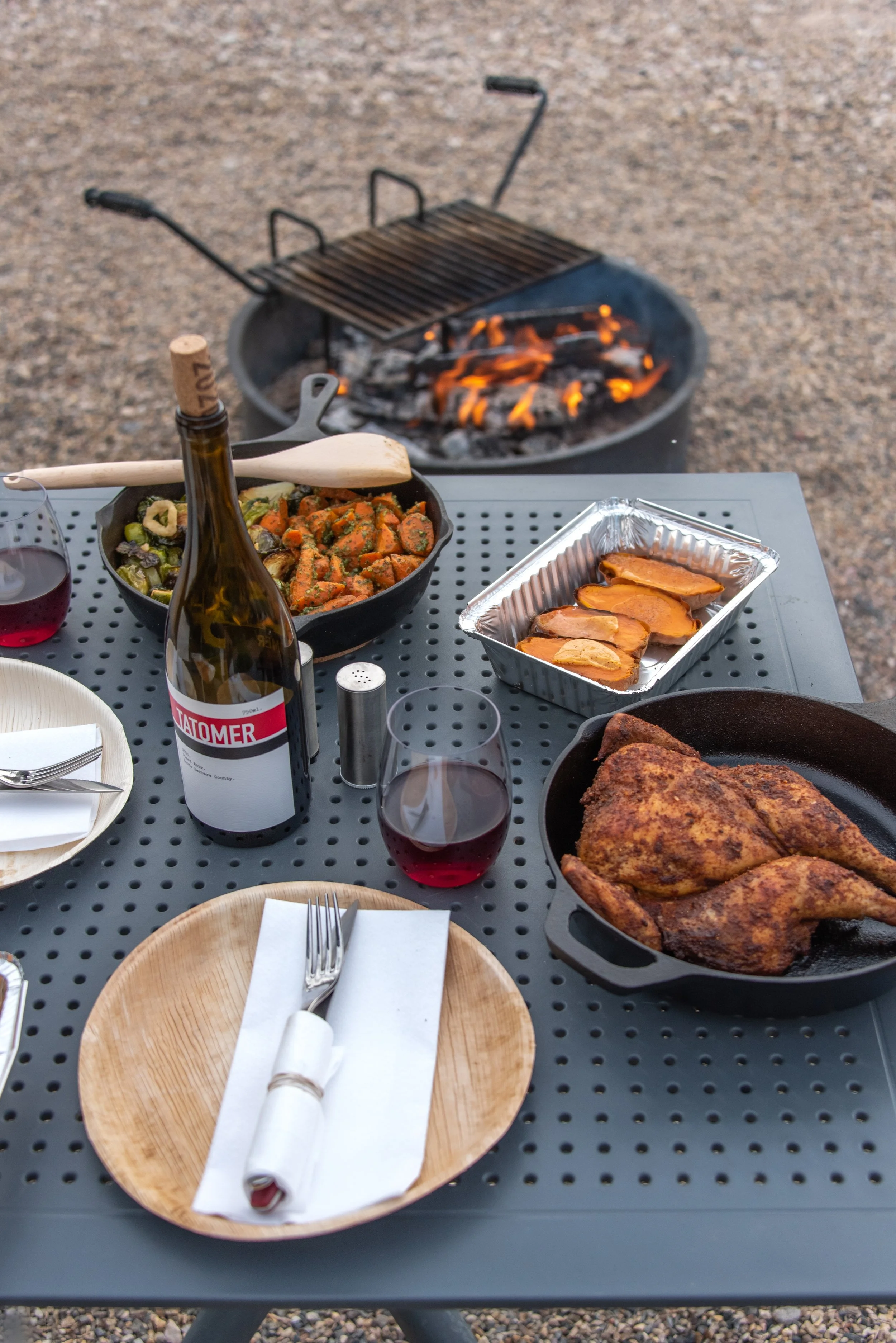 A table set outdoors with plates of roasted chicken, sweet potato slices, a bowl of roasted vegetables, and glasses of red wine, with a fire pit in the background.