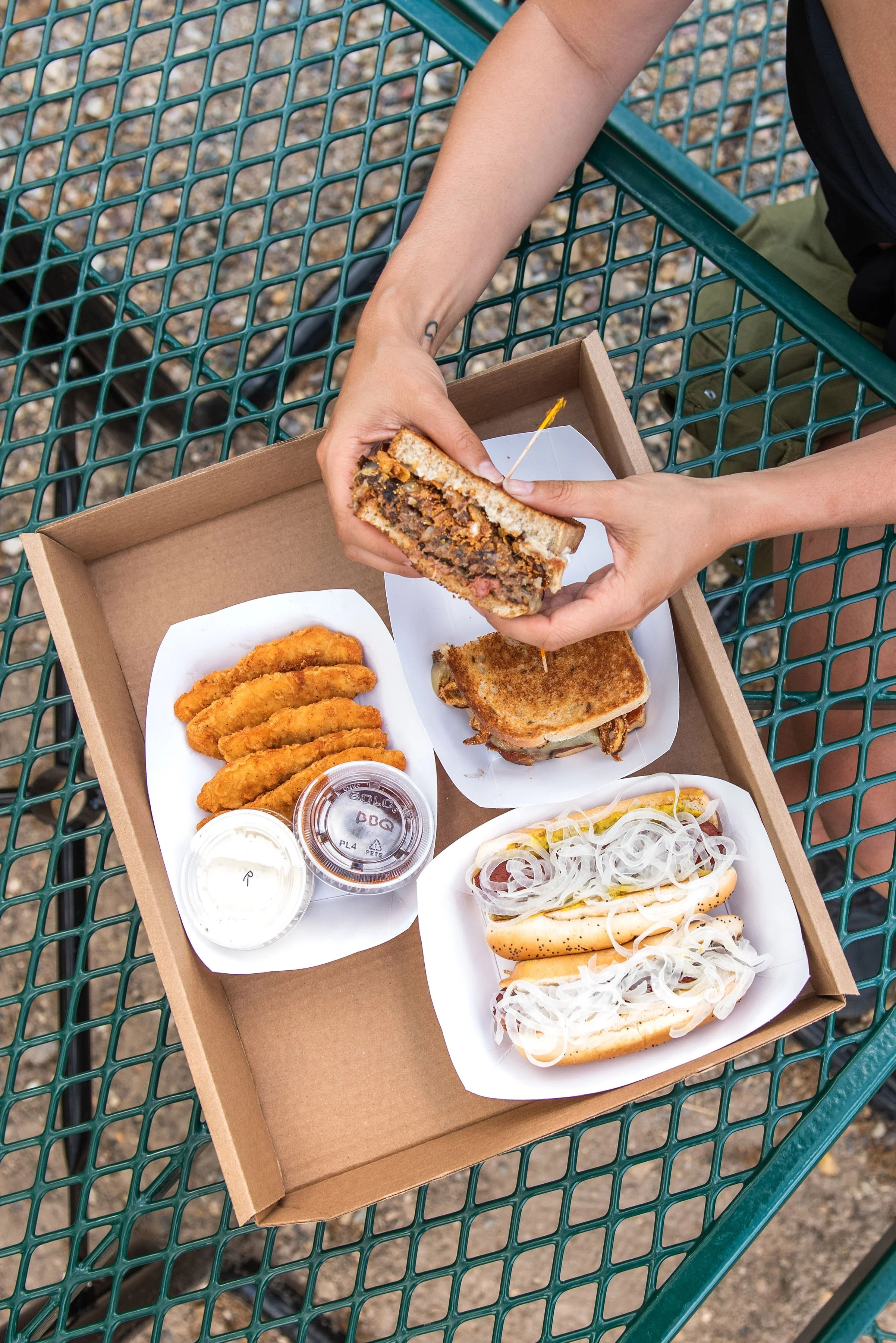 A person's hands holding half of a barbecue beef sandwich over a cardboard takeout box, on an outdoor green metal picnic table, with fried chicken tenders, onion rings, small containers of dipping sauces, and hot dogs topped with onions on white paper trays.