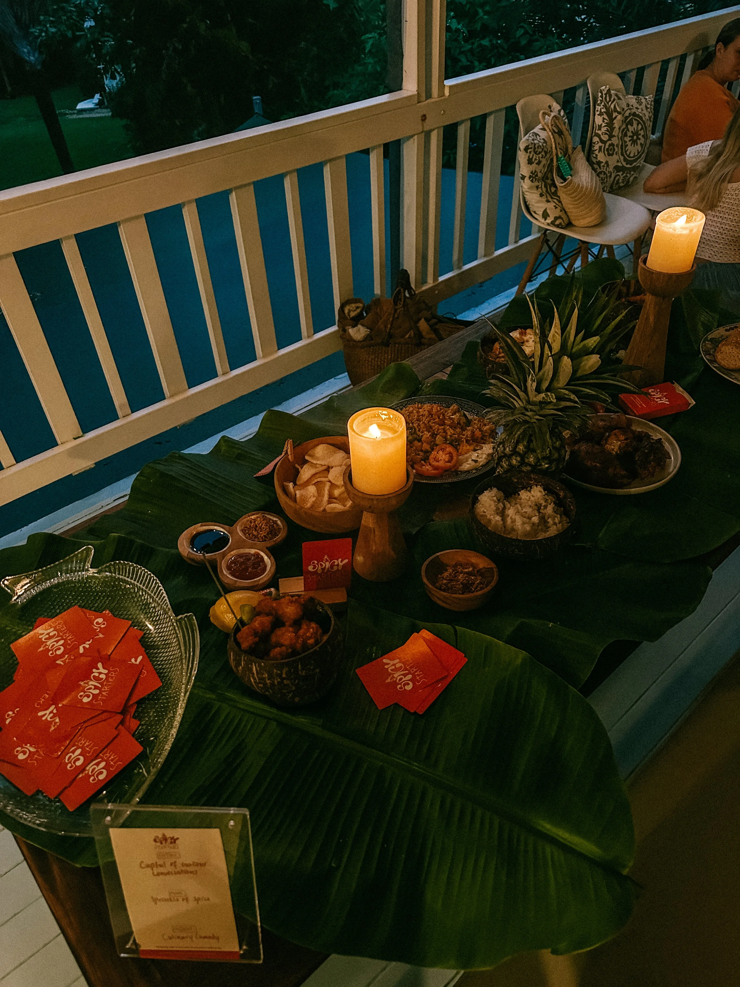 Dining table with various dishes, candles, and tropical decorations at an outdoor or semi-outdoor setting.