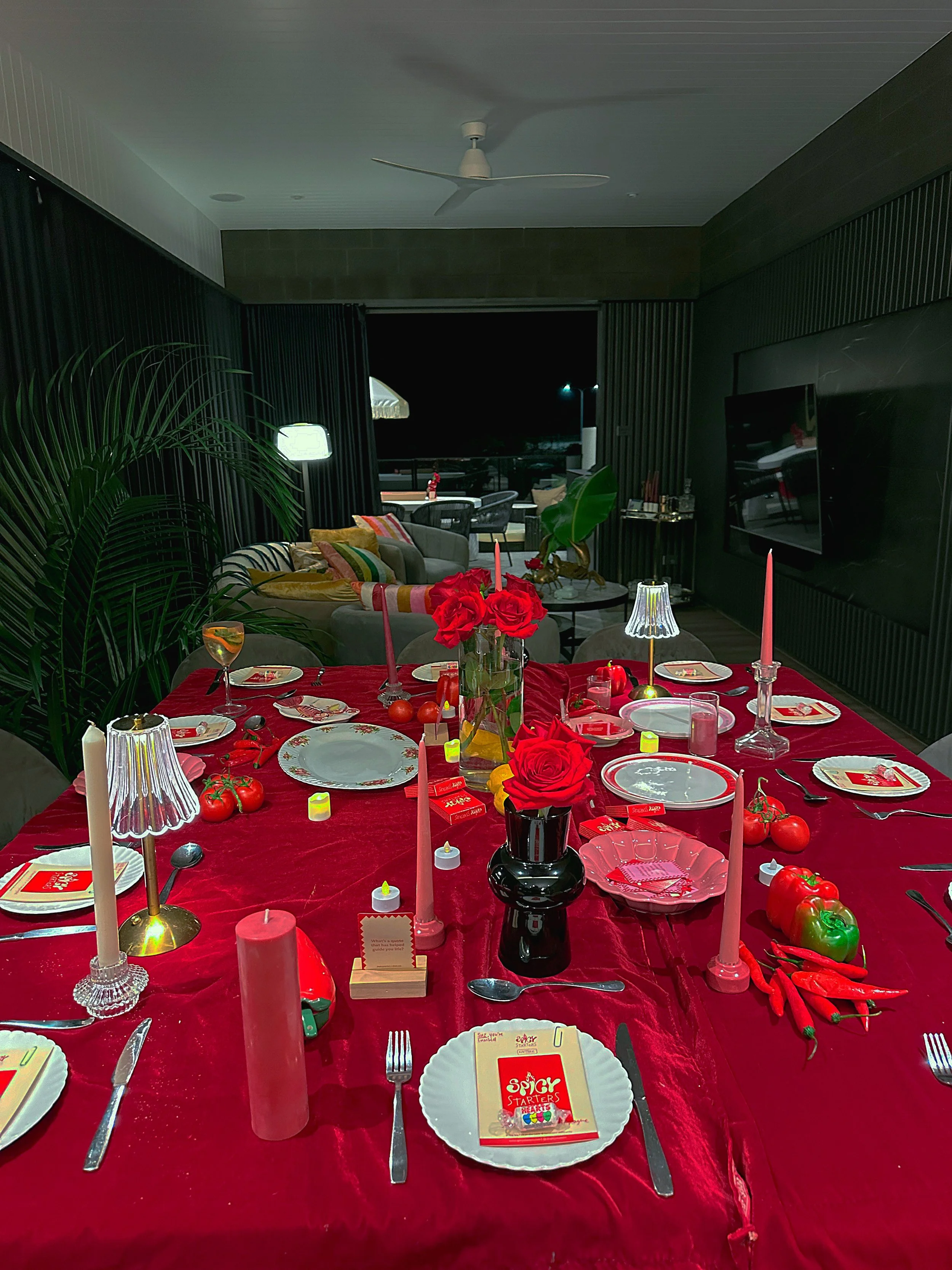 A dining table decorated for a celebration with red roses, candles, tomatoes, chili peppers, and a festive tablecloth, set with plates, silverware, and glasses.