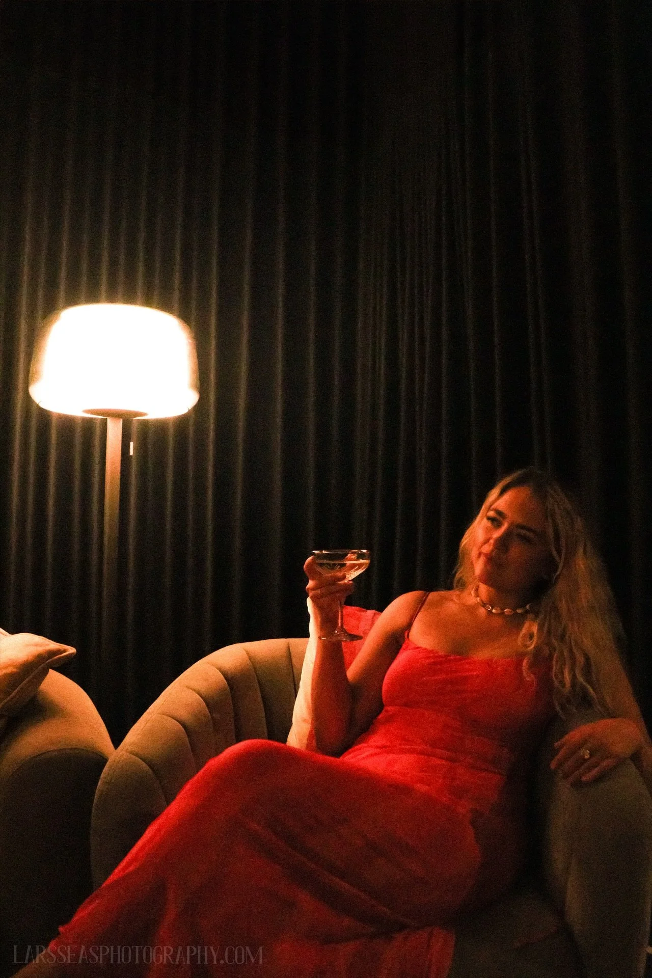 A woman in a red dress sitting on a beige armchair, holding a glass and relaxing in a dimly lit room with black curtains and a tall lamp.