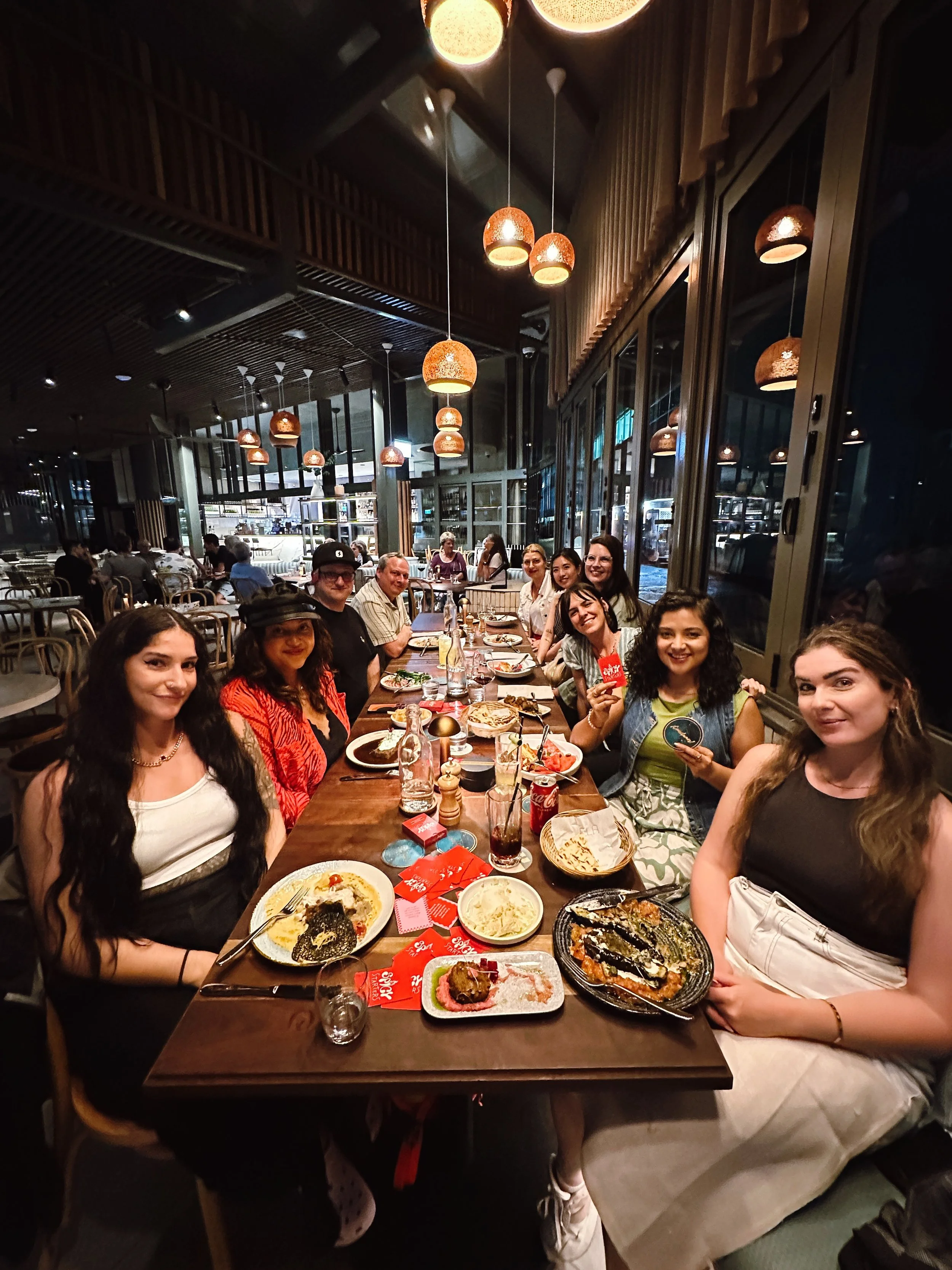 A group of people sitting at a long table in a restaurant, with various dishes and drinks in front of them, smiling and enjoying their meal.