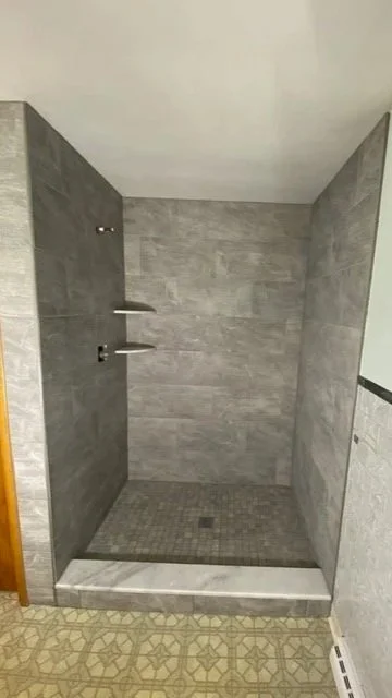 Remodeled walk-in shower with gray tile walls, a built-in shelf, and a tiled shower floor with a drain.