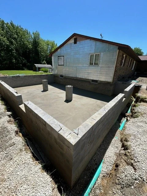 The foundation of a house under construction with concrete walls, three vertical support columns, and an attached building in the background.