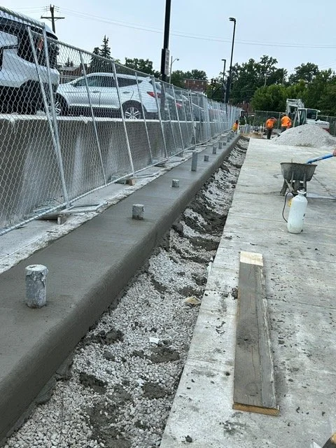 Construction worker smoothing wet concrete sidewalk with a trowel, next to a fence and parked cars.