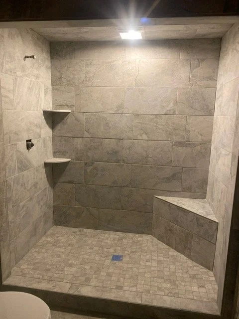 A remodeled walk-in shower with gray tiled walls and floor, featuring built-in shelves and a bench, with bright ceiling lighting.