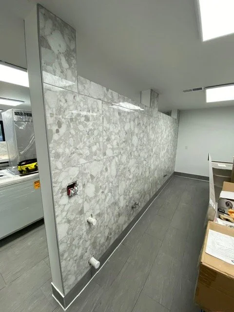 Remodeled interior room with a marble tiled wall, gray flooring, and a drop ceiling with fluorescent lights. Part of a kitchen or storage area with boxes and a counter visible on the right.