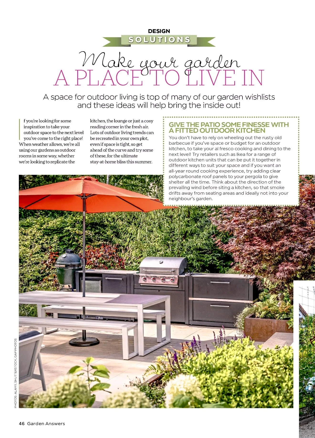 A backyard outdoor living space with a large barbecue grill, a closed grill cover, a white table, a white bench, an orange patio umbrella, and lush greenery including plants and trees.