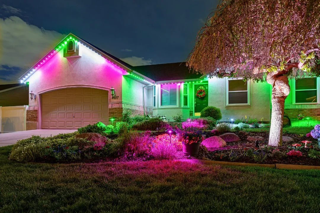 A house decorated with colorful Christmas lights at night, with pink, green, and white lights outlining the roof, a wreath on the front door, and a well-lit garden with flowers and a tree.
