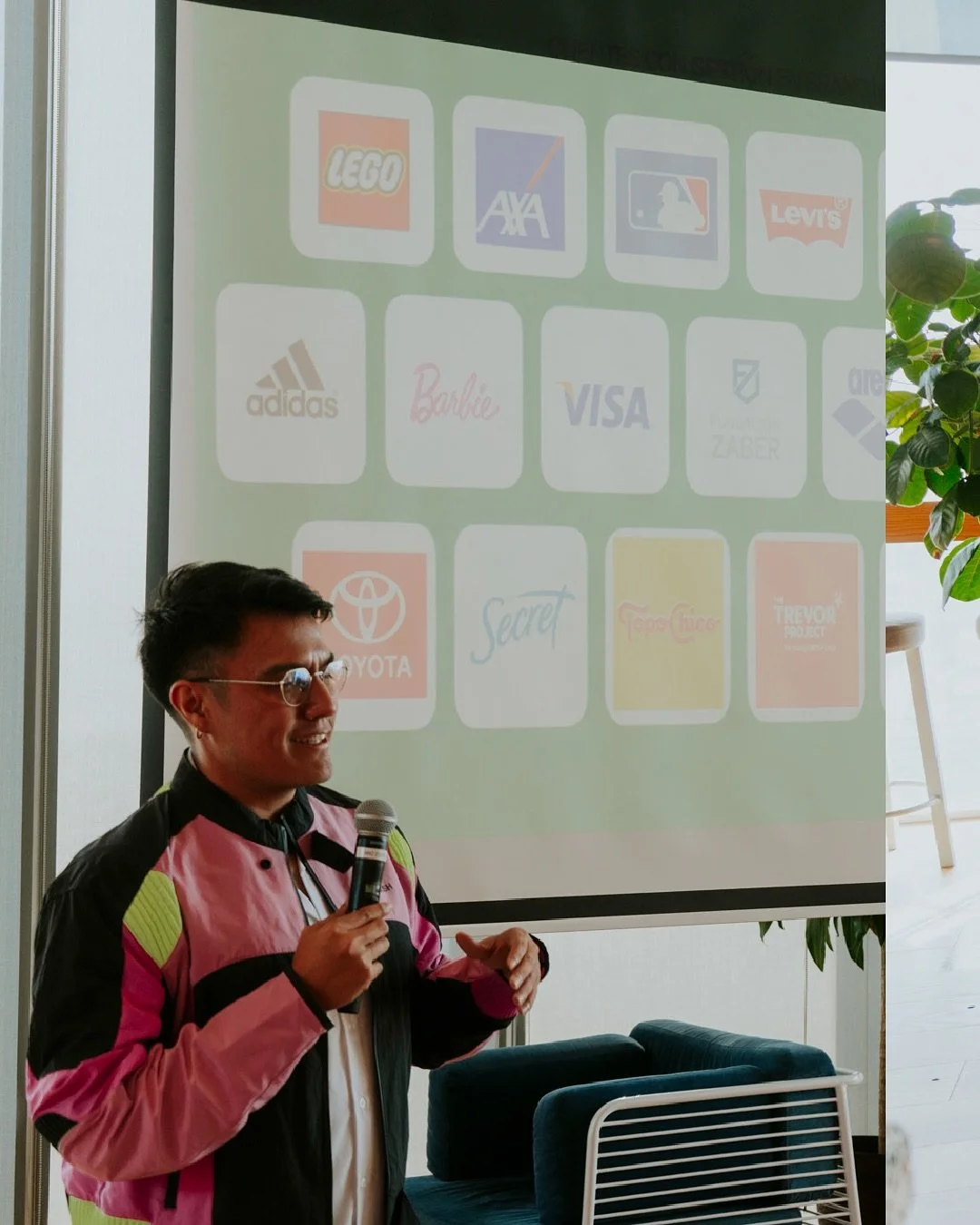 A man in a pink and black jacket holding a microphone and smiling, standing in front of a projection screen displaying various brand logos, including LEGO, AXA, Levi's, Adidas, Barbie, VISA, Toyota, and others.