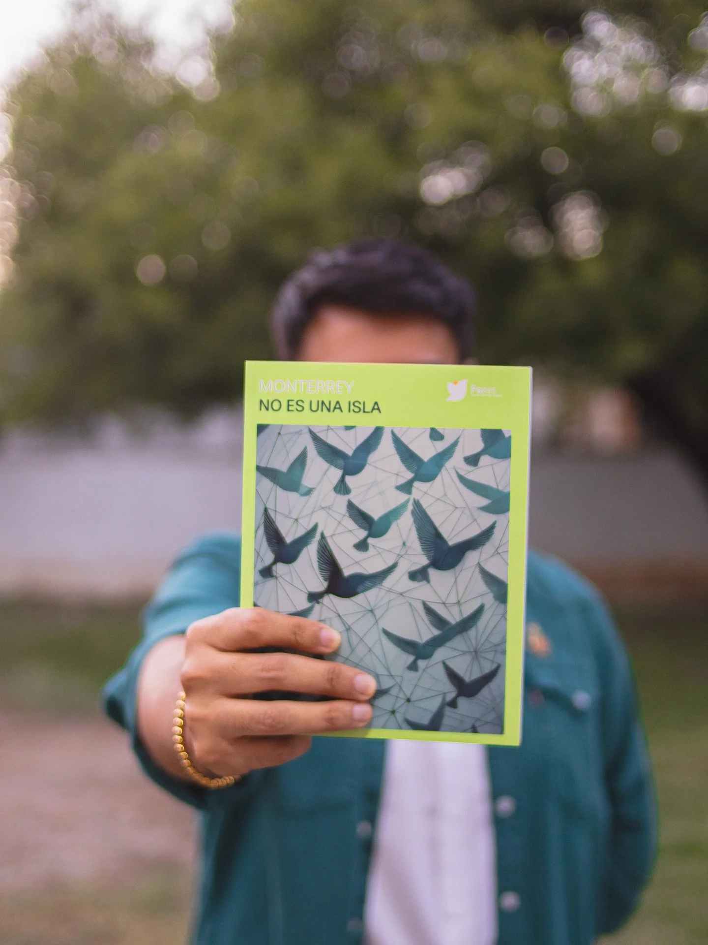 Person holding a pamphlet with illustrations of flying birds and the text "Monterrey No Es Una Isla" in front of their face. Background of trees and an outdoor setting.