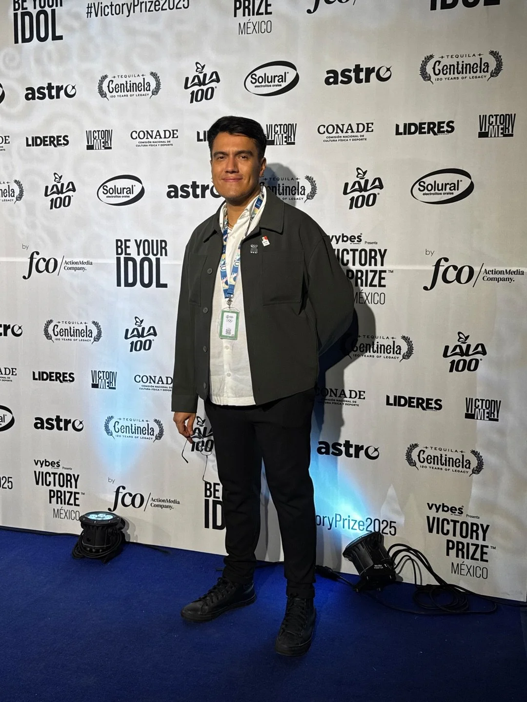 Man standing in front of a step and repeat banner with logos for Victory Prize Mexico, Astro, Tequila Centinela, La La 100, Solural, Conade, Lideres, fco ActionMedia, and Vybes. He is wearing dark pants, black shoes, a dark jacket, a white shirt, and an event badge hung around his neck.