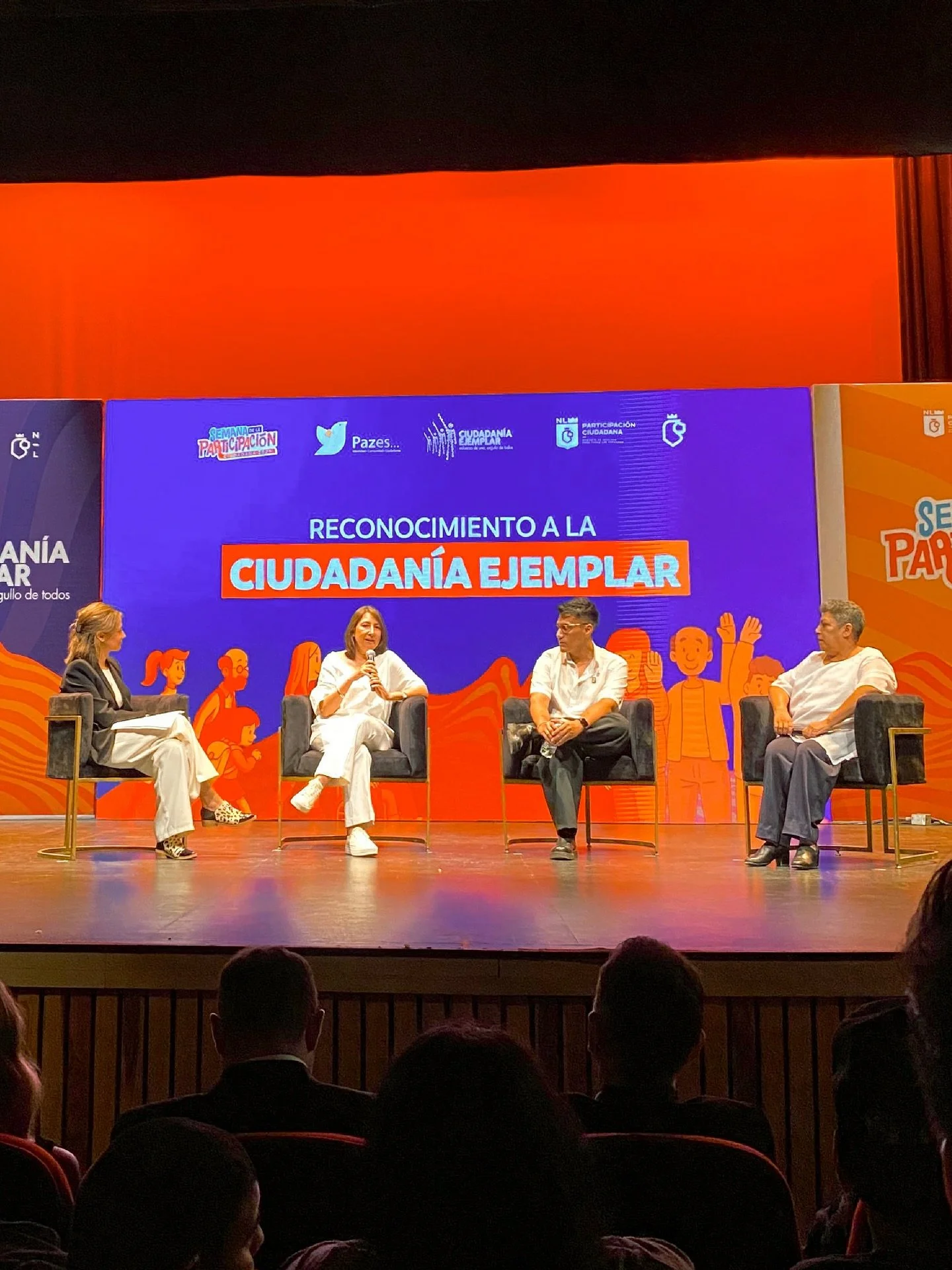 Group of four people sitting on stage in chairs during an event titled "Reconocimiento a la Ciudadanía Ejemplar," with a large blue and orange backdrop and audience members visible in the foreground.