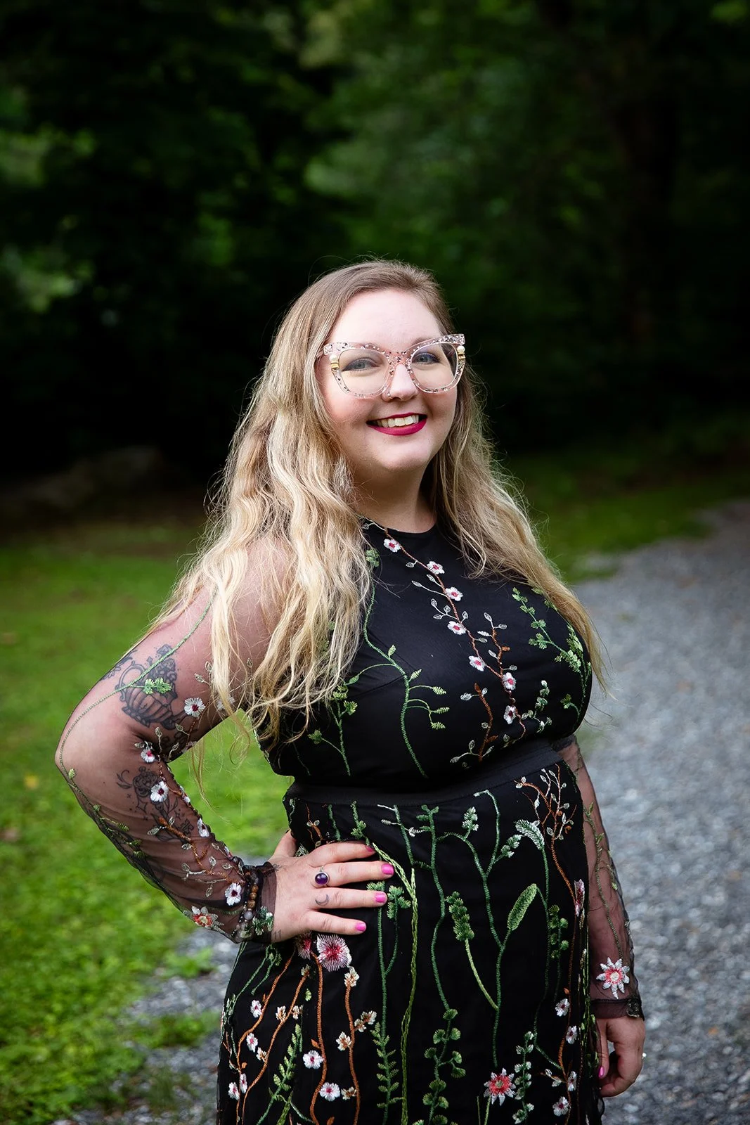 A woman with long, wavy blonde hair and glasses, smiling, standing outdoors on a gravel path with trees in the background. She is wearing a black dress with floral embroidery and sheer, embroidered sleeves, with her left hand on her hip.