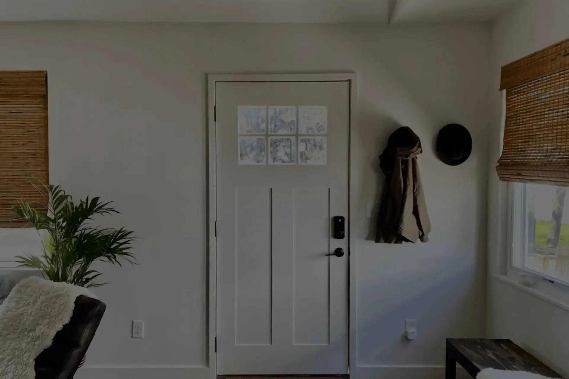 Wellness interior design entryway in Beacon New York by The Whistling Well featuring white panelled front door with glass panes wall hooks with hats and coats indoor palm plant and woven bamboo shades.webp