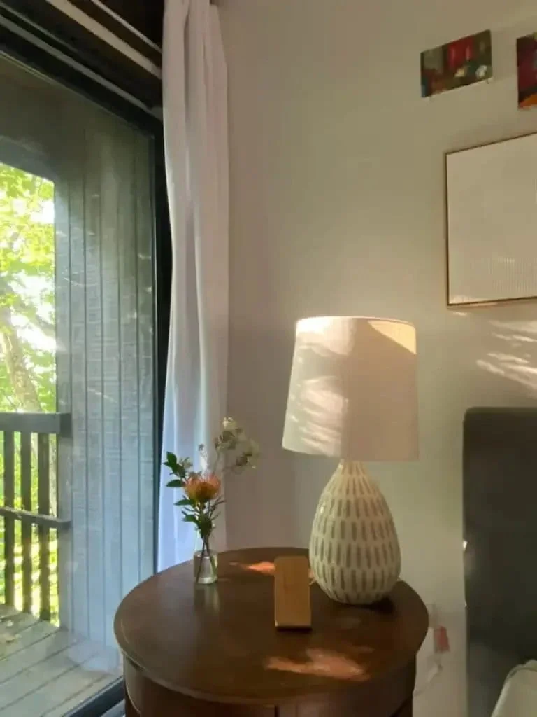 Wellness interior design bedroom detail by The Whistling Well showing walnut bedside table with ceramic table lamp single flower stem and sheer curtains with garden views.webp