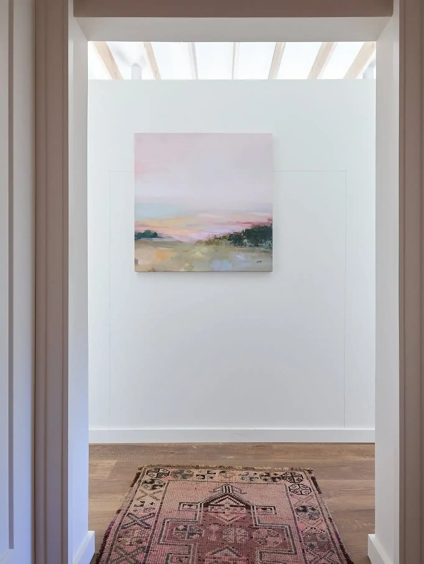 Sunlit interior with an abstract landscape painting, wood flooring, and patterned rug viewed through a doorway.