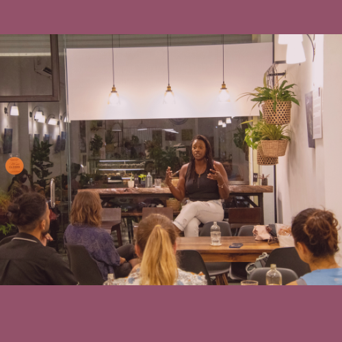 Ashleie Elaine speaking to a group in Cambodia seated around a table in a cozy, well-lit room with plants and hanging lights, during a book reading and promotion