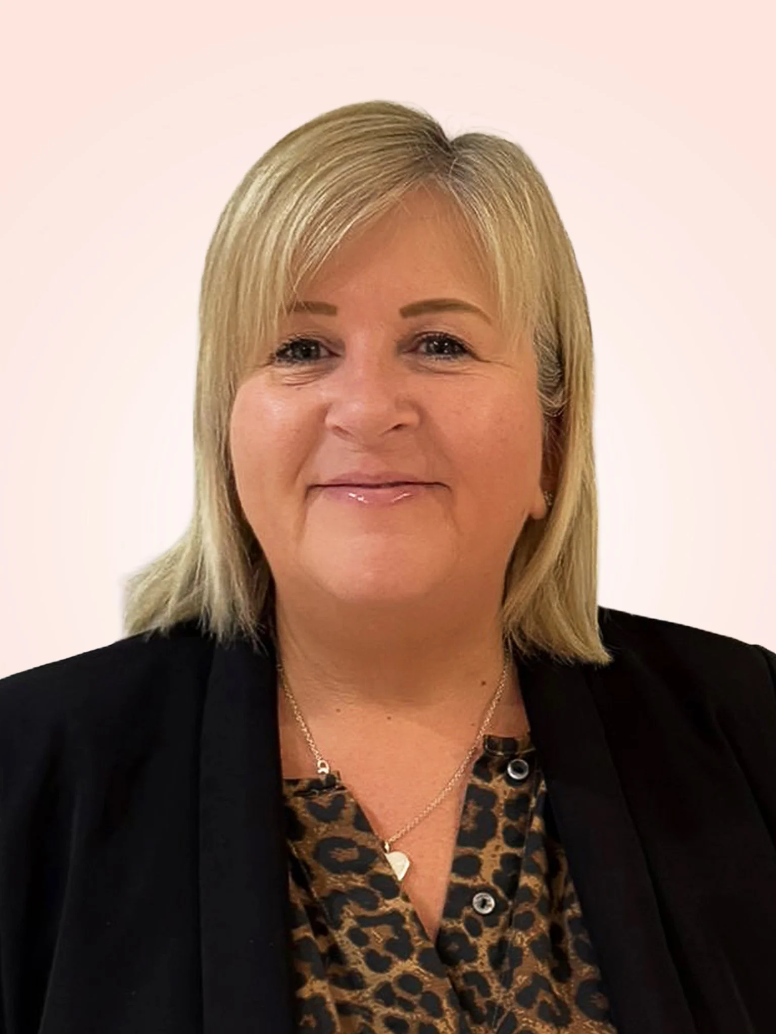 A blonde woman with shoulder-length hair wearing a black jacket, white lace top, and a black necklace, against a light pink background.