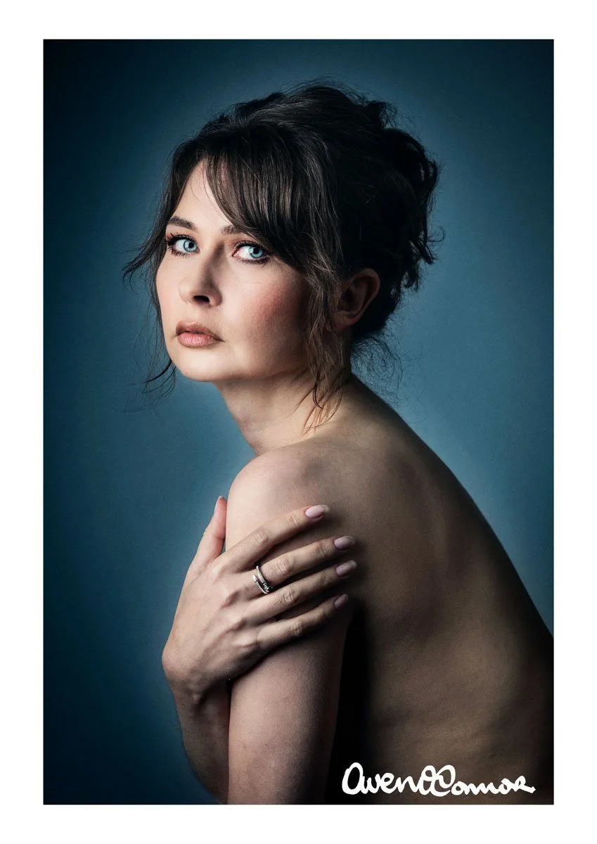A portrait of a woman with short, wavy brown hair and piercing blue eyes, standing topless with her left arm across her chest and her hand resting on her right shoulder. The background is a dark gradient, and the photograph is signed with the name "O