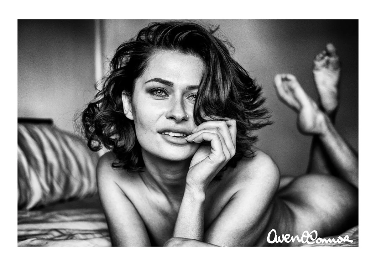 Black and white photo of a woman lying on a bed, looking at the camera, with one hand near her face, and her legs raised behind her. The photo is signed by Owen Camera.