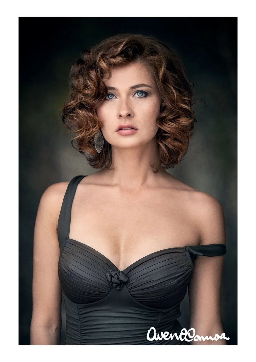 A woman with curly shoulder-length hair, blue eyes, and fair skin wearing a black, strapless dress with a small bow detail at the center, and an earring visible on her left ear.
