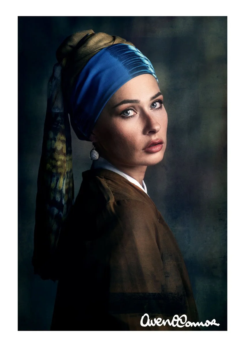A woman with fair skin and striking blue eyes, wearing a dark brown top, silver earrings, and a colorful headscarf tied up in a bun, against a dark, textured background.