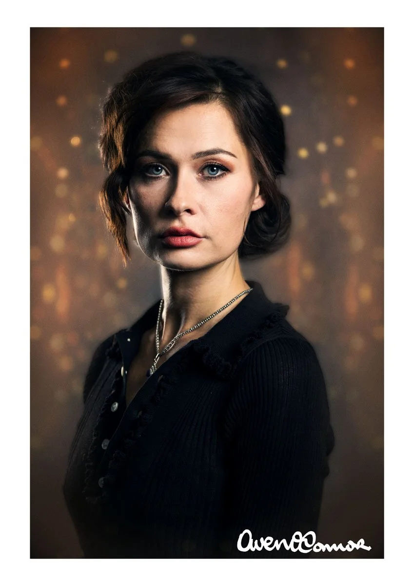 A portrait of a woman with short wavy brown hair, blue eyes, and fair skin, wearing a black top and a silver necklace, against a blurred background with warm bokeh lights.
