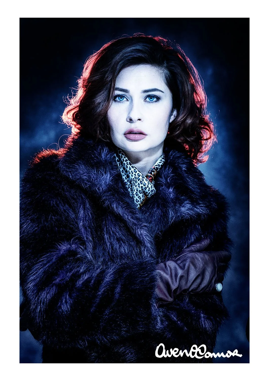 A woman with curly dark hair and blue eyes wearing a purple fur coat, a leopard print scarf, and brown gloves, posing with arms crossed against a dark background with blue and red lighting.