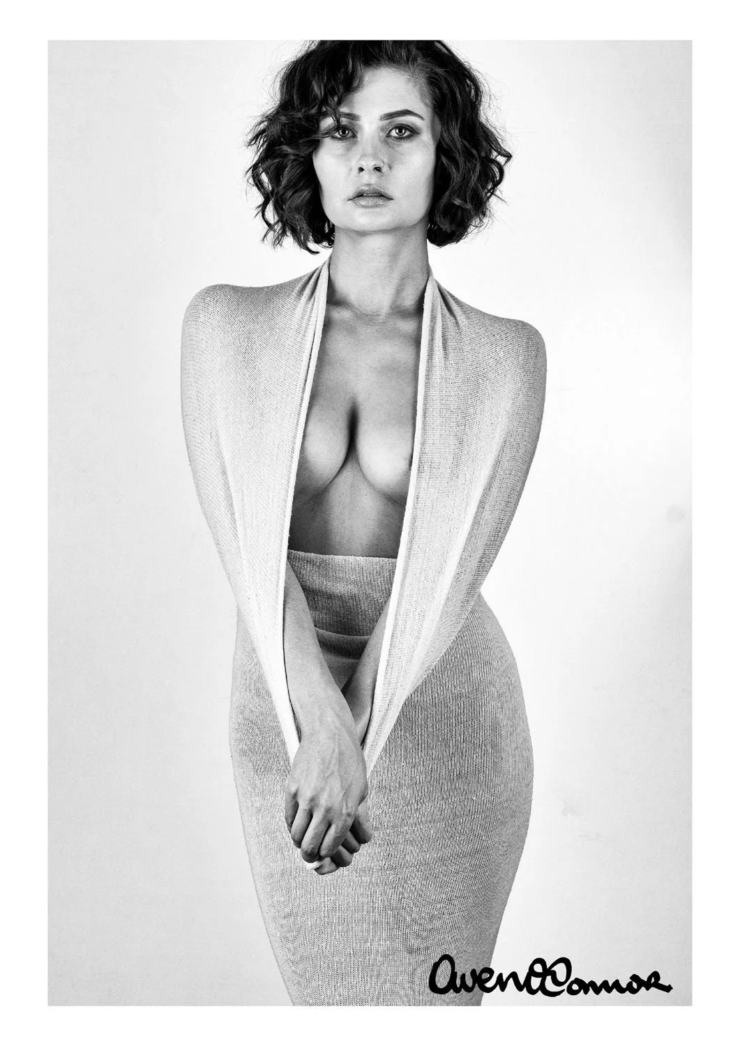 Black-and-white photo of a woman with short, curly hair wearing a light-colored knit skirt and a matching open-knit top, with her hands clasped in front of her, gazing directly at the camera. The signature 'Avenie O'Connor' appears at the bottom righ