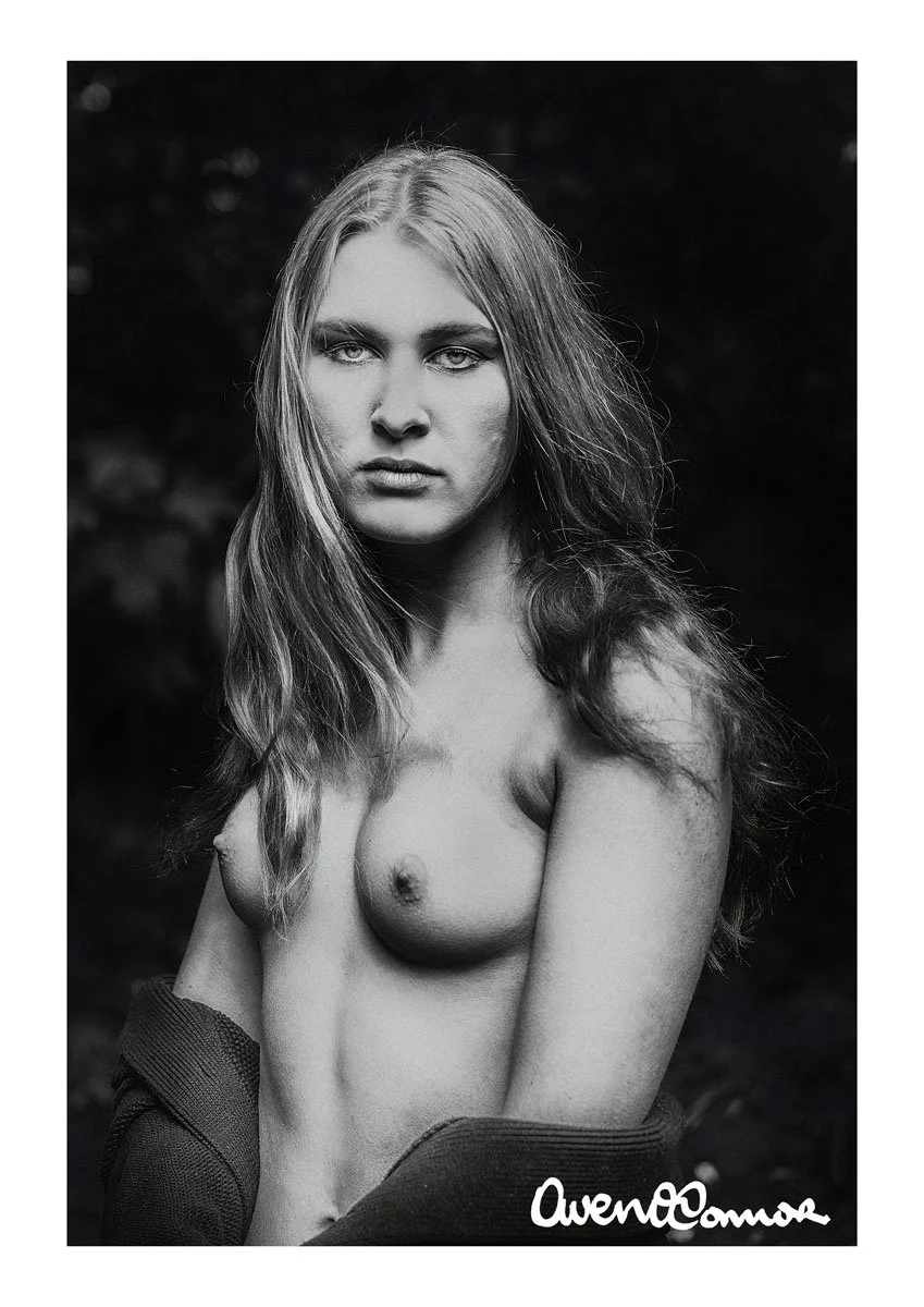 Black and white portrait of a topless woman with long wavy hair, looking directly at the camera, with a dark blurred background and an artist signature in the bottom right corner.