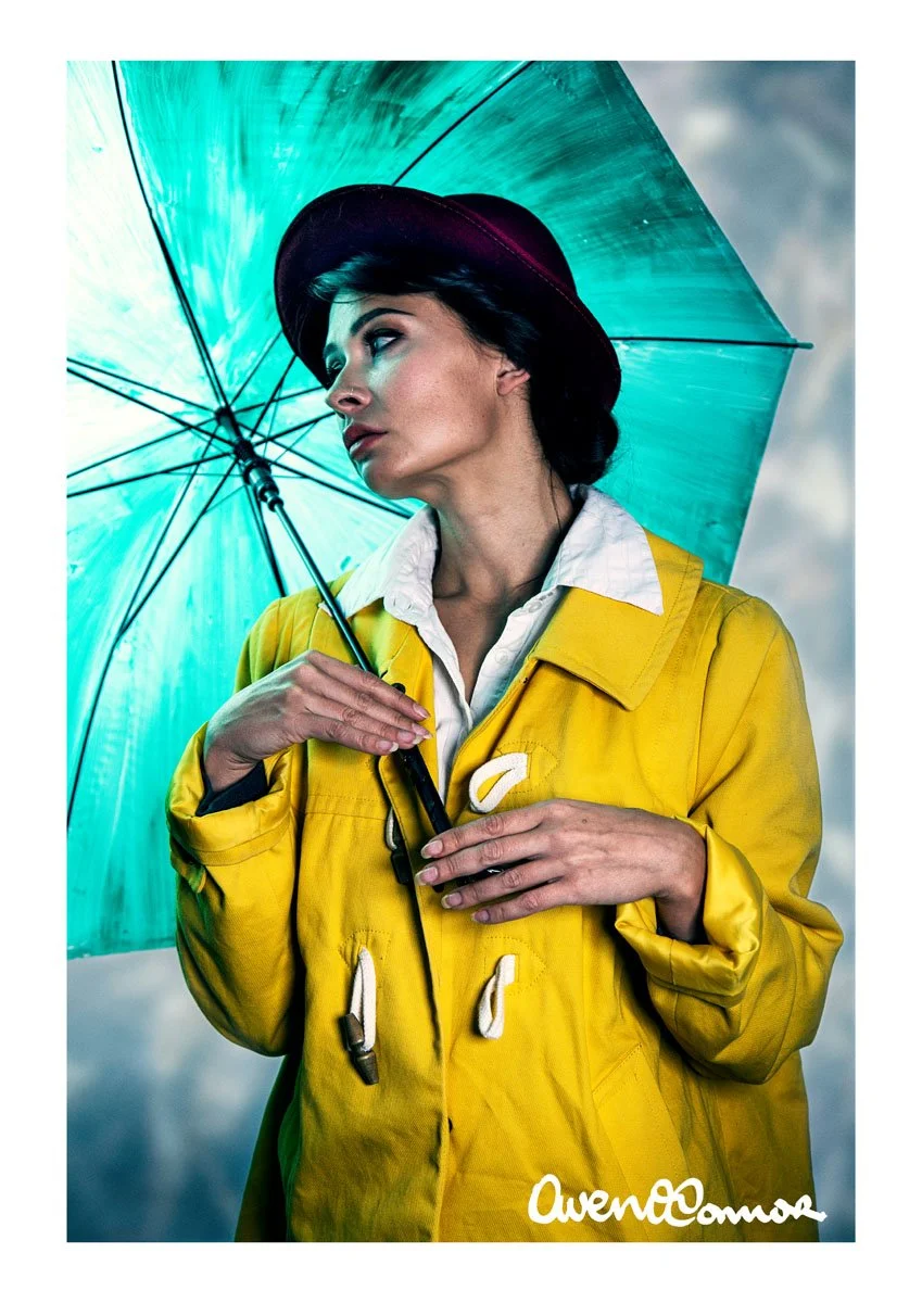 A woman with dark hair wearing a yellow raincoat, white shirt, and black hat holding a turquoise umbrella, standing outdoors against a cloudy sky.