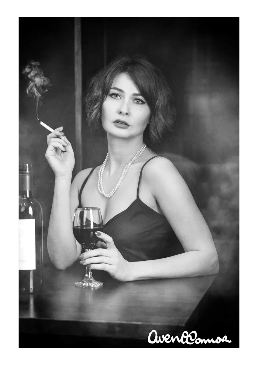 A black-and-white photograph of a woman with short, wavy hair, wearing a spaghetti strap dress and pearl necklace. She is holding a glass of red wine in one hand and a lit cigarette in the other, with smoke rising from it. She is looking directly at