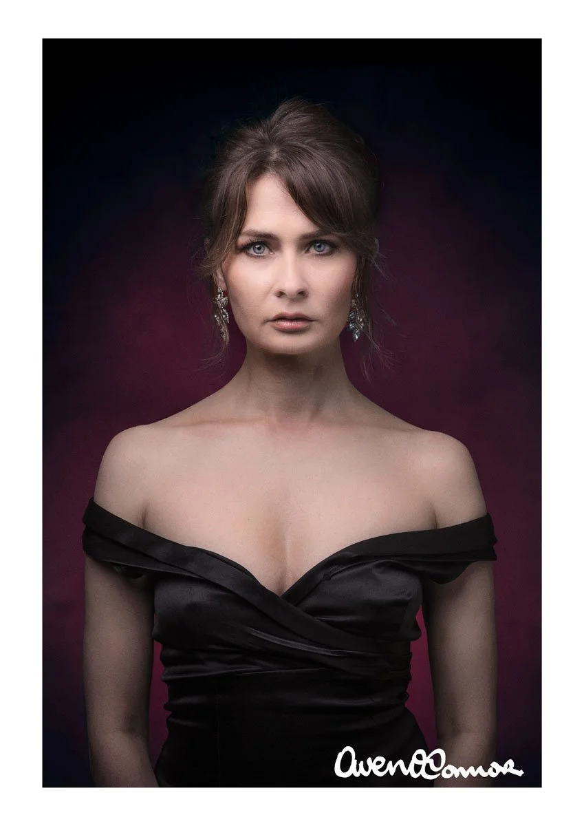 A woman with brown hair styled in an updo, wearing dark eyeshadow and earrings, in a black off-the-shoulder dress, against a purple and pink gradient background, looking directly at the camera.