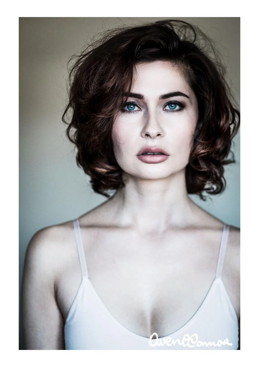 Close-up portrait of a woman with curly brown hair and bright blue eyes, wearing a white spaghetti strap top, against a neutral background.