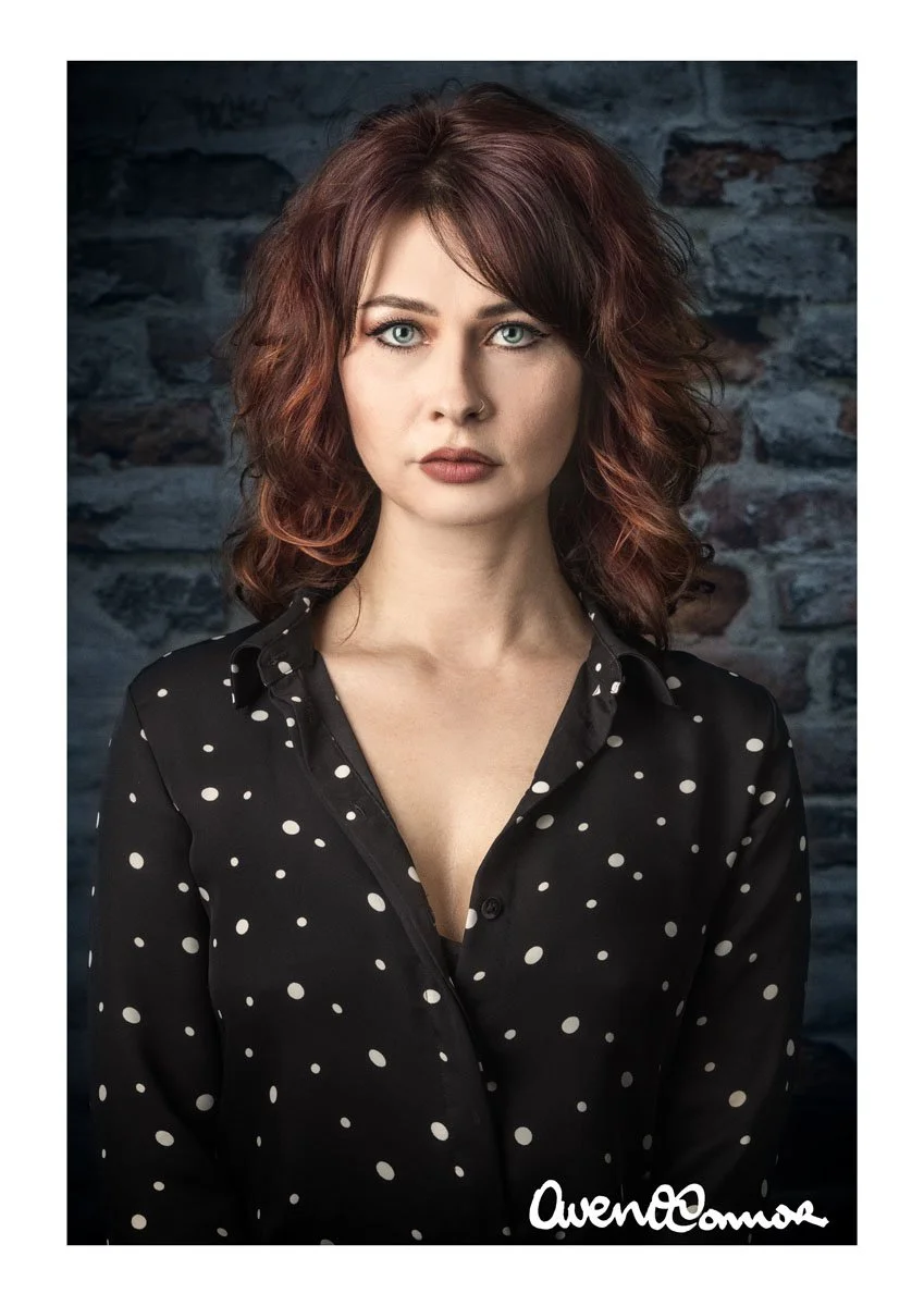 A woman with wavy red hair and blue eyes looks directly at the camera, standing against a dark stone wall background. She is wearing a black shirt with white polka dots, partially unbuttoned.