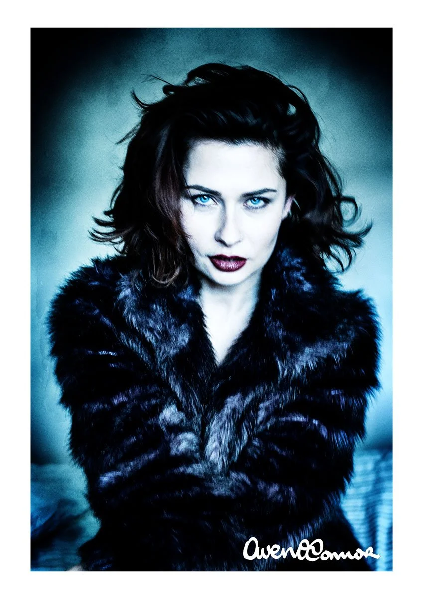 A woman with dark, curly hair and striking blue eyes poses with her arms crossed, wearing a black fur coat against a dark, muted background.
