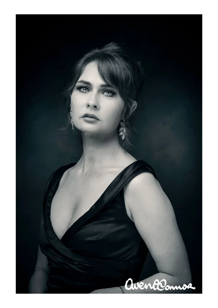 Black and white portrait of a woman with wavy hair wearing earrings and a satin dress with a plunging neckline, against a dark background.