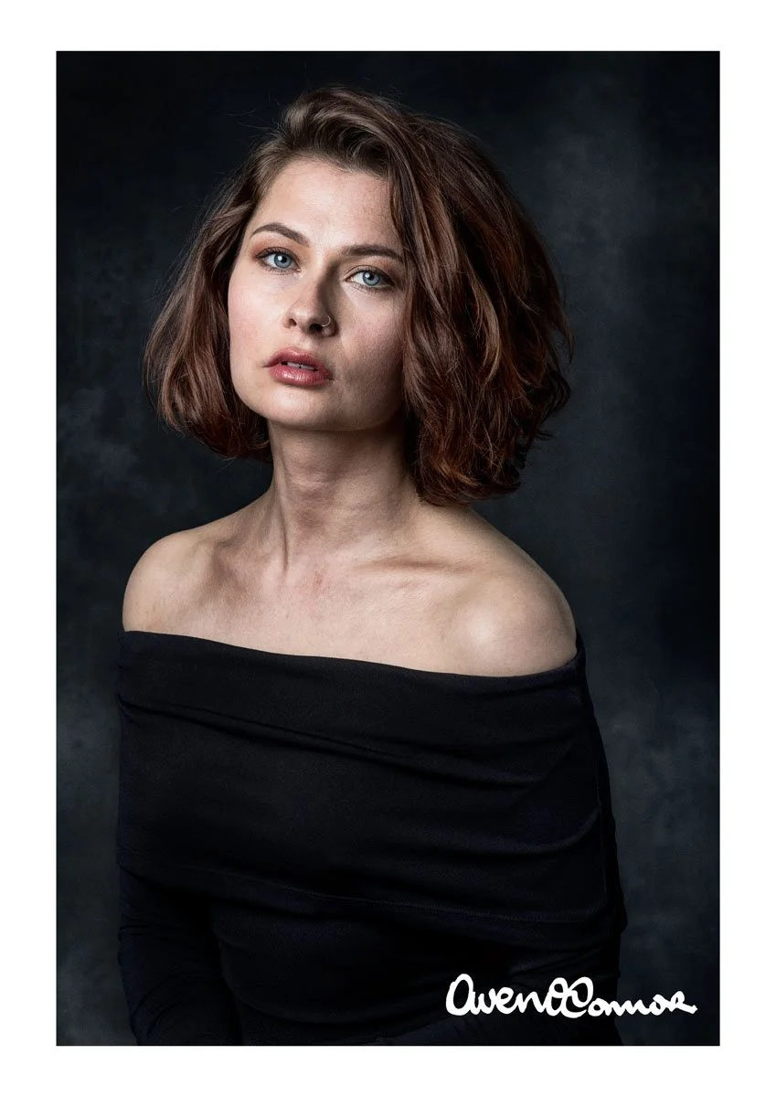 A woman with wavy auburn hair, blue eyes, and a nose piercing, wearing an off-the-shoulder black top against a dark background.