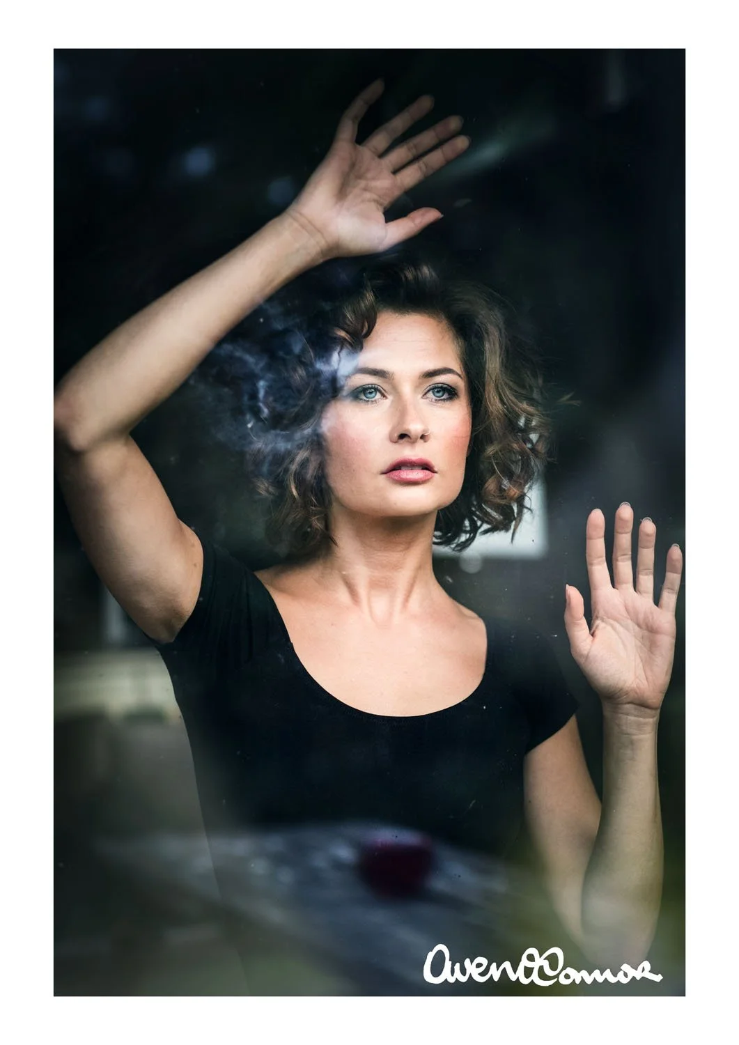 Woman with curly hair in a black shirt standing behind a glass window, looking thoughtfully outside with her hands on the glass.
