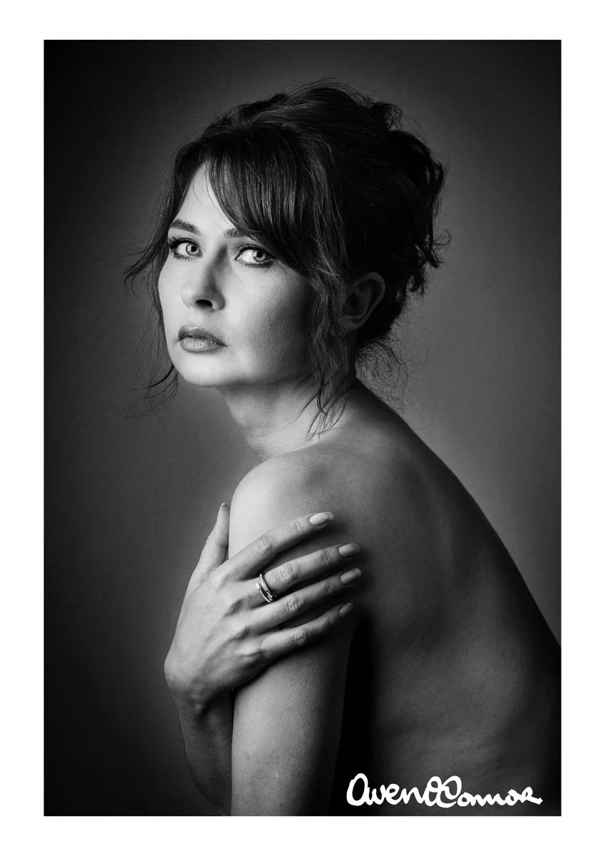 Black and white portrait of a woman with short, wavy hair, gazing at the camera, with her hand touching her shoulder, wearing a ring on her finger.