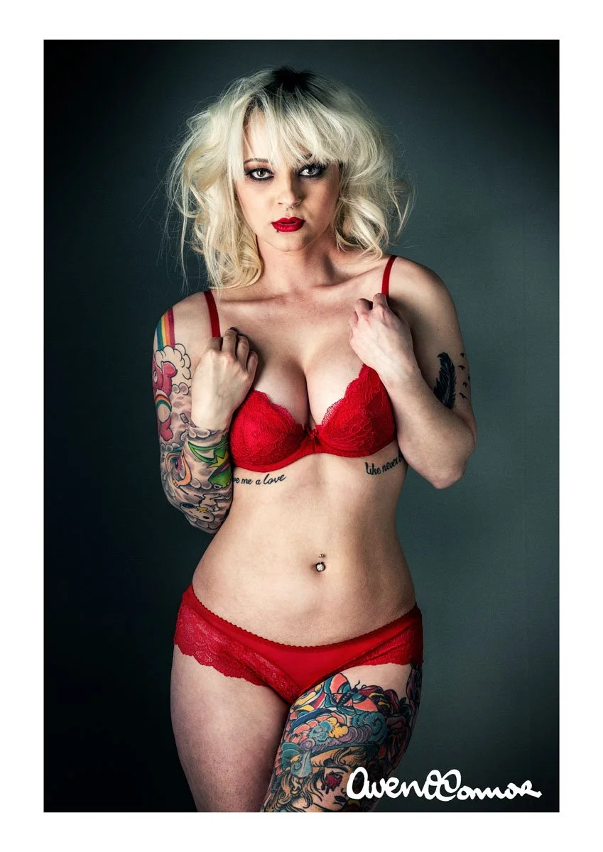 A tattooed woman with platinum blonde hair and red lipstick wearing red lace lingerie poses against a dark background.