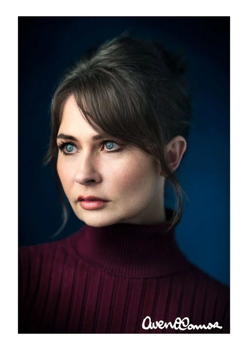 Close-up portrait of a woman with blue eyes and brown hair in a bun, wearing a burgundy turtleneck sweater, against a dark background.