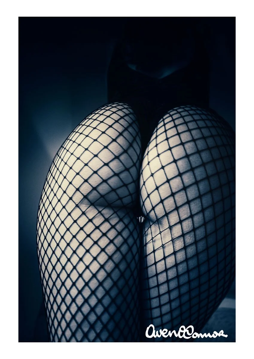 Close-up of a woman wearing fishnet stockings, with her knees bent and her body partially obscured in shadow.